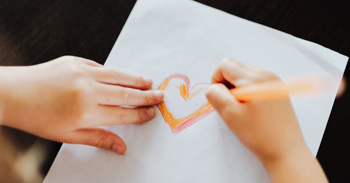 A child drawing a heart with colored pencils on white paper, emphasizing creativity and innocence.